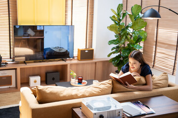 Stay home pastimes. Asian woman sitting in cozy sofa couch with open book indoors. Beautiful young lady reading book, enjoying lazy morning, having relaxing weekend.