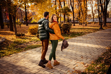 Fototapeta premium Smiling couple walking in an autumn park under sunshine, surrounded by golden foliage, enjoying a lovely bonding moment