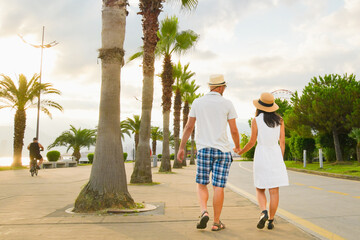 Young couple tourists walking hand in hand along Batumi promenade with palm trees and seaside view on sunrise, concept of tourism, vacation, lifestyle and romantic travel in Georgia