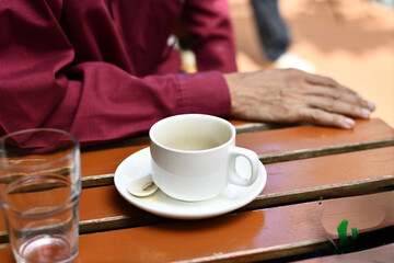 White coffee cup on saucer with used spoon on wooden table near elderly hand in red shirt conveying quiet morning contemplation
