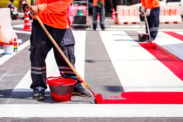 road worker applying fresh red paint to a crosswalk stripe with roller and bucket during street maintenance under bright sunlight
