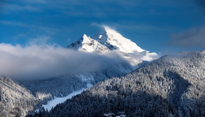 A Snow Capped Mountain Peak Partially Obscured By Clouds Surrounded By A Forested Mountainous Landscape Under A Blue Sky