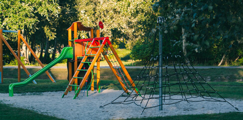 Empty playground with colorful slide, climbing frame, and rope pyramid in a park. Outdoor recreational area for children surrounded by grass and trees on a sunny day.