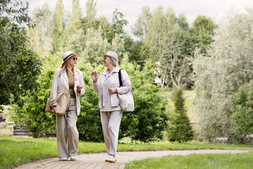 Fototapeta premium Full length portrait of two senior women walking outdoors in stylish light beige and cream casual outfits with hats, linen trousers, jackets, bag and fashionable accessories in natural park background