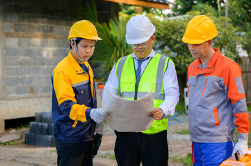 Three Asian builders examine a house that is still under construction. As the team points out roof details, the foreman holds rolled blueprints. Workers have on helmets and vests.