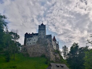 Bran Castle, located in Romania, stands prominently atop a rocky hill. The medieval fortress features a mix of gray stone and red-tiled roofs with tall, crenelated towers. Dracula's legend