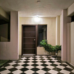 Illuminated, modern entrance dark wooden door with metal strips, a window, and a potted plant. Black and white checkered floor.