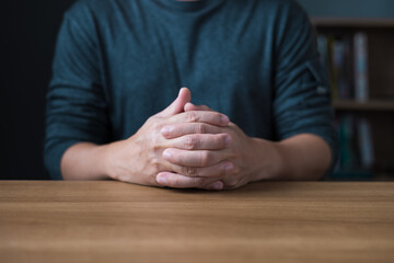 A person sits at a wooden table with hands clasped together,conveying feelings of waiting,patience,focus,anticipation,decision making,calm presence,thoughtfulness,and professional body language.