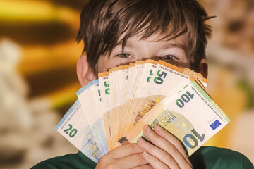 Smiling boy proudly holding a fan of euro banknotes in 20, 50, and 100 denominations, symbolizing wealth, savings, finance, and prosperity.