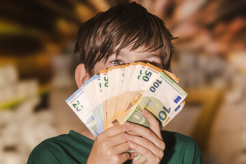 Smiling boy proudly holding a fan of euro banknotes in 20, 50, and 100 denominations, symbolizing wealth, savings, finance, and prosperity.