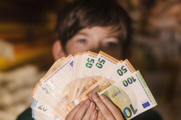 Smiling boy proudly holding a fan of euro banknotes in 20, 50, and 100 denominations, symbolizing wealth, savings, finance, and prosperity.
