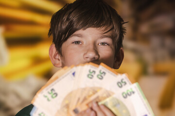 Smiling boy proudly holding a fan of euro banknotes in 20, 50, and 100 denominations, symbolizing wealth, savings, finance, and prosperity.