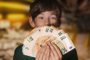 Smiling boy proudly holding a fan of euro banknotes in 20, 50, and 100 denominations, symbolizing wealth, savings, finance, and prosperity.