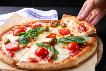 Woman taking piece of delicious pizza with cheese, tomatoes and olives at dark table, closeup