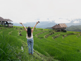 A joyful woman stands on rice terraces, raising her arms in freedom while holding a camera, with...