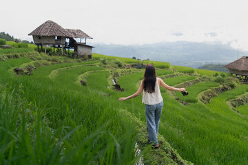 A woman walking joyfully through green rice terraces, arms open wide, wooden hut and misty...
