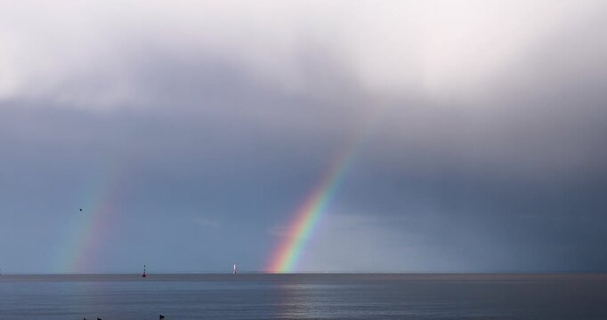 Double rainbow over water in real time. The arcs drift as rain curtains move across the scene. Secondary arc visible. 30 fps, no people.