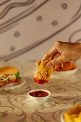 A hand holds a crispy chicken piece above a small bowl of ketchup, with a burger and other food items visible in the background.