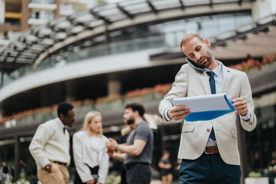 A businessman diligently works while talking on the phone and reviewing documents outdoors. In the background, colleagues are seen interacting near a modern corporate building. - Powered by Adobe