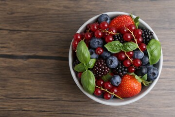 Different ripe berries and basil leaves in bowl on wooden table, top view. Space for text