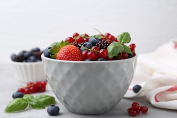 Different ripe berries and basil leaves in bowl on light grey table, closeup