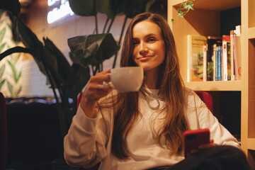 Brunette long hair woman use mobile phone while sitting in comfortable coffee shop during evening, charming happy female reading news, social media, answer on message, laughing or smiling.