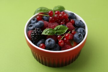 Different ripe berries and basil leaves in bowl on green background, closeup