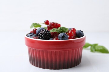 Different ripe berries and basil leaves in bowl on white tiled table, closeup