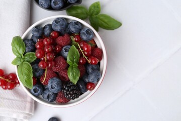 Different ripe berries and basil leaves in bowl on white tiled table, flat lay. Space for text