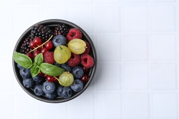 Different ripe juicy berries and basil in bowl on white tiled table, top view. Space for text