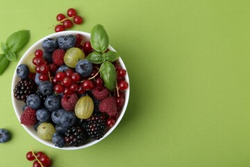 Different ripe juicy berries and basil in bowl on green background, flat lay. Space for text