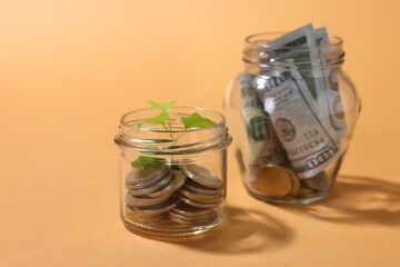 Glass jars with coins, sprout and dollars on pale orange background, closeup