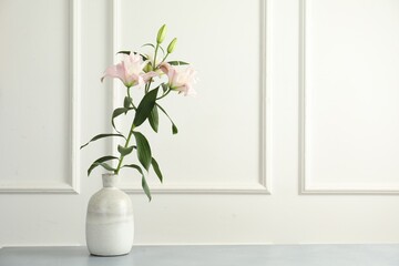 Beautiful lily flowers in vase on grey table near white wall, space for text
