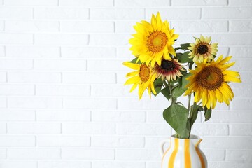 Beautiful sunflowers in jug against white brick wall indoors, space for text