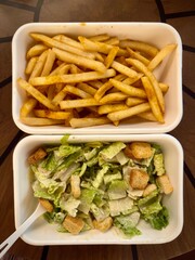 Caesar salad and french fries in white takeout containers on wooden table
