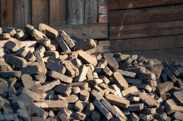 A large pile of coal bricks is arranged next to a weathered wooden wall, illuminated by warm afternoon sun