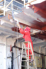 Repairman on ladder is spraying primer white color on steel ceiling inside of engine room at the stern of oil tanker ship