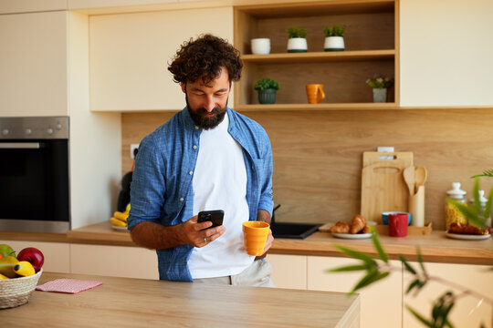 Smiling man drinks coffee while checking phone in a bright modern kitchen - Powered by Adobe