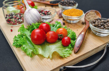 Cherry tomatoes, garlic, dry spices in bowls and spoons on  wooden cutting board on black backdrop.