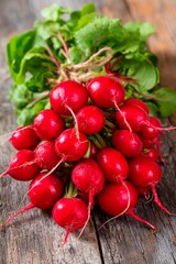 A bunch of vibrant red radishes is neatly gathered with green leaves attached against a rustic wooden background. The fresh vegetables are ready for use in cooking