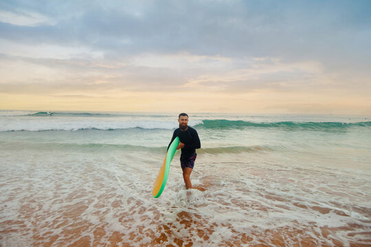 Smiling young man with surfboard on the beach. Boy coming out of the ocean after water surfing.