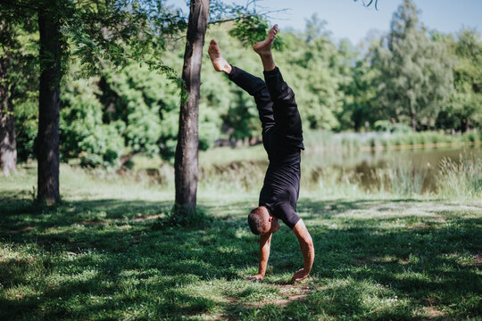 A person executes a handstand in an open, grassy park area with trees and sunlight. The tranquil outdoor setting highlights the enjoyment of fitness and nature together.