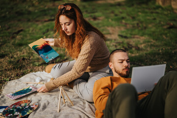 A couple relaxes outdoors on a blanket, engaging in artistic pursuits like painting.