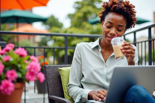 Analyzing Data on Laptop: African American Woman Enjoys Fancy Iced Coffee on Colorful Rooftop Patio with Modern Decor and Blooming Flowers