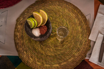 Overhead shot of a delicious, crystal-clear mezcal in its glass caliber, accompanied by worm salt, oranges, and lemon served in a small molcajete.

