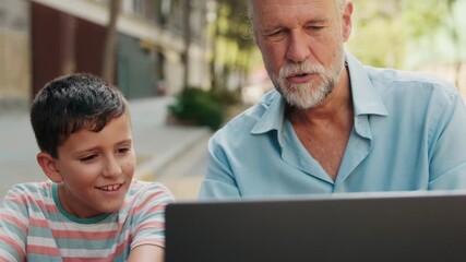 Grandfather and grandson sit together at a table outdoors, looking at a laptop and smiling. They are enjoying their time together.
