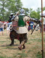 Two armored fighters in medieval combat reenactment. Participants wear full helmets, shields, and weapons while engaging in battle during a historical reenactment festival outdoors.
