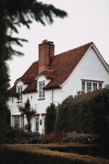 Fototapeta premium A charming white cottage adorned with a vibrant red roof rests serenely among lush greenery, creating a picturesque scene in this tranquil rural landscape on a cloudy day