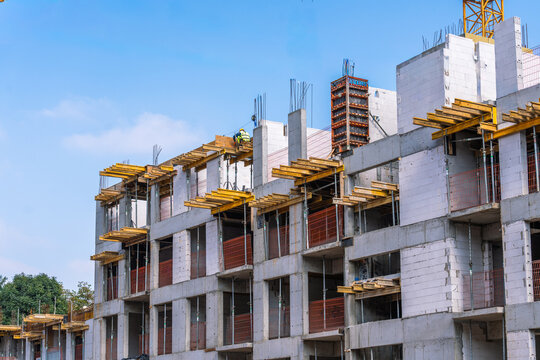 Construction site of a residential building with concrete framework and wooden supports against a blue sky backdrop. Workers present.