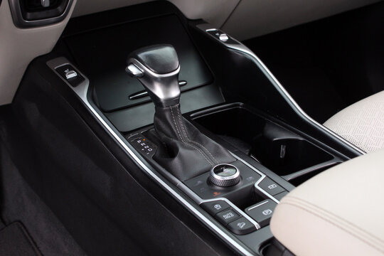 Modern car center console with automatic gear shift lever in black leather, chrome accents, control buttons, and rotary dial, surrounded by beige and black car interior.